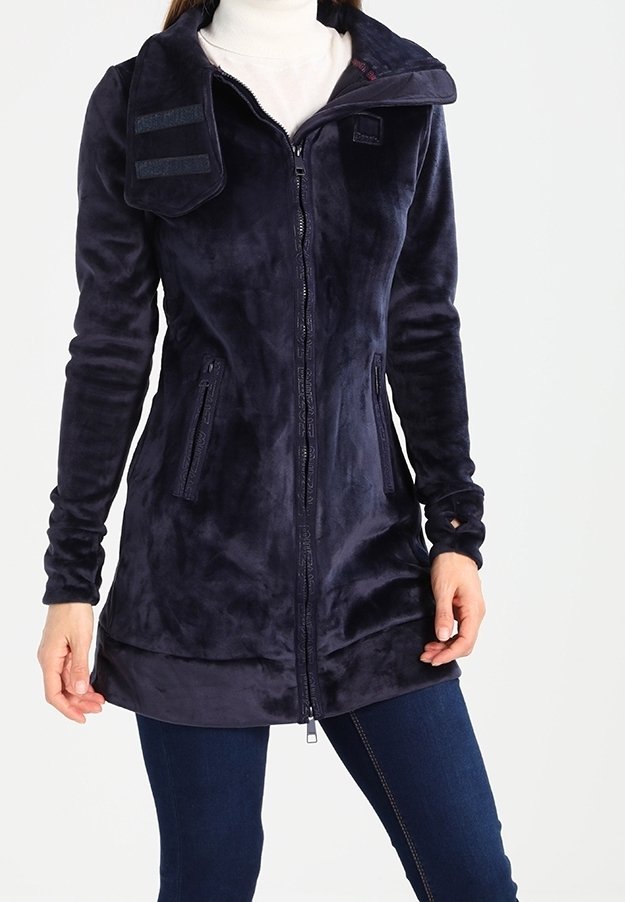 Donna che indossa una lunga giacca in fleece blu navy con zip, dotata di un colletto alto e tasche laterali con zip, abbinata a jeans blu scuro.