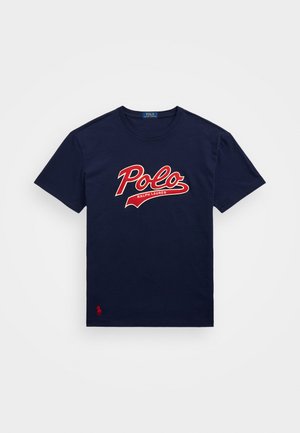 Tamsiai mėlynas medvilninis marškinėliai su dideliu raudonu "Polo" logotipu su baltomis detalėmis, trumpomis rankovėmis ir mažu raudonu logotipu apatinėje kairėje pusėje.