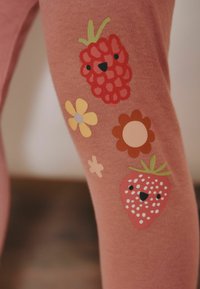 Vaaleanpunaiset legginsit, joissa on leikkisiä kuvioita mansikoista, vadelmista ja kukista eri väreissä, ja niissä on sarjakuvamaisia ilmeitä.