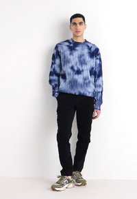 Modra in bela tie-dye kapuca s sproščenim fitom, v kombinaciji s črnimi slim-fit hlačami in zeleno/sivimi športnimi čevlji.