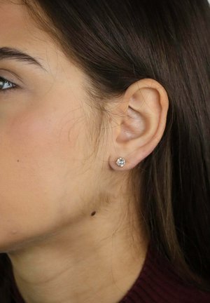 Gros plan sur l'oreille gauche d'une femme aux cheveux brun foncé, portant une petite boucle d'oreille en diamant ronde.