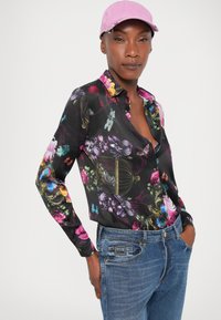 Camicia nera con motivo floreale, decorata con farfalle colorate e rose, maniche lunghe e colletto. Abbinata a jeans in denim azzurro chiaro e un cappellino rosa.