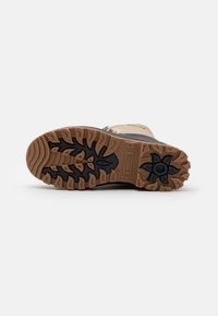 Semelle en caoutchouc marron avec un motif de traction texturé noir. La chaussure possède un dessus en matériau beige avec une accentuation foncée sur le côté.