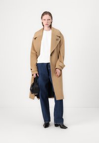 Cappotto in lana cammello con ampi revers, chiusura a bottoni, abbinato a una camicia bianca e jeans a vita alta blu scuro, e una piccola borsa nera.