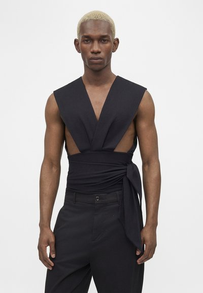 Άνδρας με κοντά ξανθά μαλλιά, που φοράει αμάνικο μαύρο τοπ τύπου wrap και μαύρο παντελόνι μπροστά σε απλό ανοιχτό φόντο.