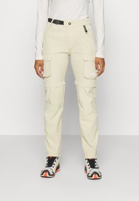 Ljusbeige cargobyxor tillverkade av ett slitstarkt material, med ett bälte, två stora sidofickor, logotyp på låret och en tapered passform.