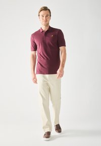 Bordeaux polo skjorte med korte ærmer og klassisk krave, parret med lyse beige bukser og brune sko. Diskret logo detalje på skjorten.
