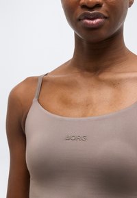 Κοντινό πλάνο ενός ατόμου που φοράει τοπ ταουπ με λεπτές τιράντες και τη λέξη «BORG» ανάγλυφη στο στήθος.