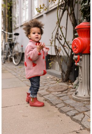 Ružový flísový kabát s červenými motívmi jahôd, červeným lemovaním a gombíkmi. Tmavé riflové nohavice a červené gumáky. Mestské prostredie s bicyklom a hydrantom.