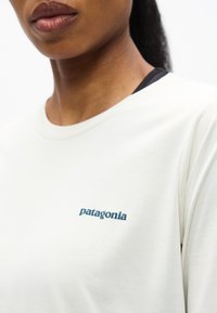 Κοντινό πλάνο ατόμου που φοράει λευκό πουκάμισο Patagonia με ορατό λογότυπο στο στήθος και μερικώς εμφανή χείλη και λαιμό.