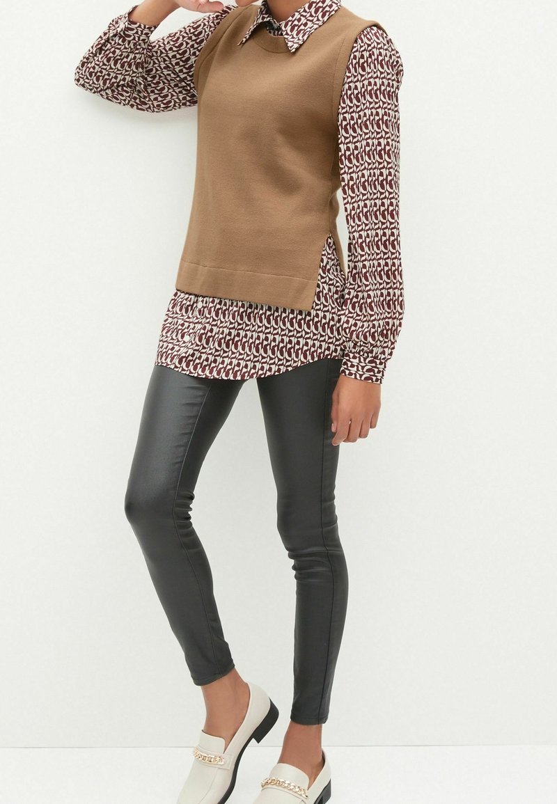 Brauner ärmelloser Pullover über einer gemusterten Bluse mit einem weißen und kastanienbraunen Design, kombiniert mit schwarzen Leggings aus Kunstleder und weißen Slippern.