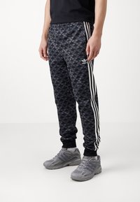 Svartmönstrade joggingbyxor med ett subtilt lövmönster, vita sidostrippar, elastisk midja och muddade anklar, kombinerade med grå sneakers.