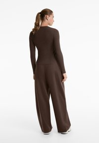 Donna con coda di cavallo che indossa una maglia aderente marrone a maniche lunghe e pantaloni larghi marroni a gamba larga, in piedi di spalle su uno sfondo bianco.