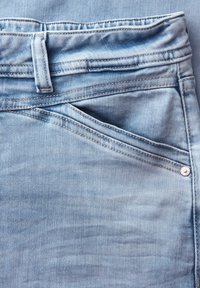 Primo piano di jeans in denim blu chiaro che mostra la tasca anteriore, il passante per cintura e i dettagli delle cuciture visibili.