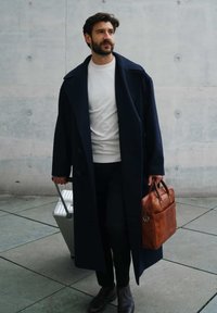 Navy overcoat, white shirt, black pants, brown leather bag, silver suitcase on textured floor against a gray wall. Smooth fabric and structured design.
