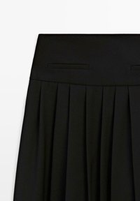 Jupe noire plissée avec une large ceinture et deux poches fendues horizontales près du haut, présentée sur un fond blanc.