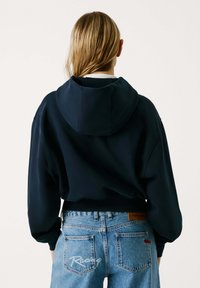 Mornarsko plava dukserica s kapuljačom i opuštenim krojem, s nisko postavljenim ramenima i rebrastim manžetama, u kombinaciji sa svijetloplavim trapericama.