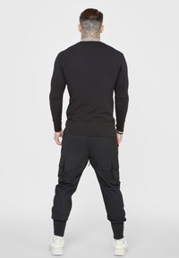 Muž s podrobným tetováním na krku, oblečený v černém tričku s dlouhým rukávem, černých cargo kalhotách a bílých teniskách, stojící zády na neutrálním pozadí.