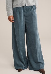 Person i brede blå fløjl bukser, grå strikket cardigan og brune spidse sko, står med den ene hånd i lommen.