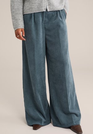 Persoon in brede blauwe corduroy broek, grijze gebreide cardigan en bruine puntige schoenen, staand met één hand in de zak.