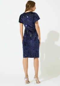 Navyblaues Pailletten-Kleid mit kurzen Ärmeln und taillierter Silhouette, das einen knielangen Saum und eine glatte Textur aufweist. Silbernene Absätze akzentuieren den Look.
