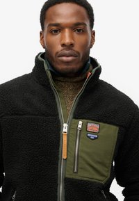 Zwarte fleecejas met olijfgroen schouderpaneel, ritszakken en logo's. Heeft een hoge kraag en een getextureerd materiaal.