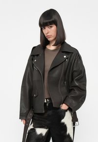 Brown leather biker jacket with silver hardware, zippered pockets, a wide collar, and side belt detail, worn over a brown top and patterned pants.