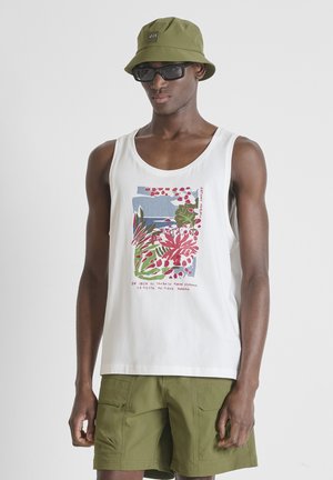 Man draagt een olijfgroene bucket hat, zwarte zonnebril, wit tanktop met kleurrijke abstracte print en olijfgroene cargoshorts, staand tegen een grijze achtergrond.