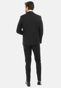 Completo nero con un taglio su misura, caratterizzato da uno spacco singolo sul retro. Il materiale sembra liscio, abbinato a eleganti scarpe nere lucide.