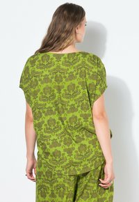 Top verde a fantasia con un design floreale, maniche corte e larghe, e orlo arrotondato. Il materiale appare leggero e strutturato.