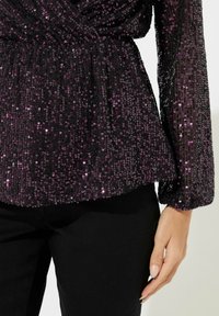 Zwarte top met paarse pailletten, voorzien van opgezette lange mouwen en geplooid middel, gecombineerd met zwarte broek met een gestructureerd oppervlak.