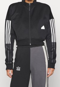 Svart zip-up kort jacka med vit tre-stripesdesign på ärmarna och en liten vit logotyp på bröstet, tillverkad av ett slätt material.