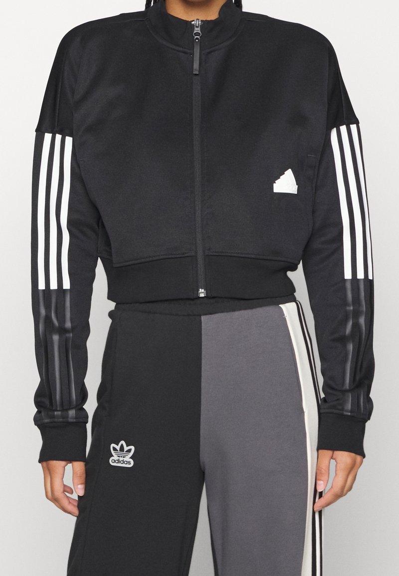 Svart zip-up kort jacka med vit tre-stripesdesign på ärmarna och en liten vit logotyp på bröstet, tillverkad av ett slätt material.