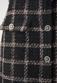 Tissu noir texturé avec un motif à carreaux comprenant des accents dorés et roses. Cinq boutons en argent ornés d'un design d'étoile décorent le devant.