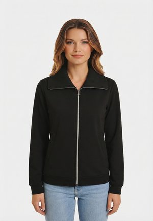 Mujer con chaqueta negra con cremallera y cuello ancho acanalado, y vaqueros azules claros, de pie frente a un fondo blanco liso.