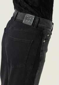Schwarze Denim-Jeans mit einer glatten Textur, Knopfverschluss und einem markanten rechteckigen Logo-Patch am Bund.