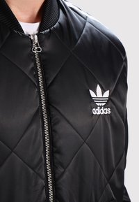 Chaqueta bomber negra acolchada con cuello de canalé, que presenta un logo bordado en blanco contrastante. El zipper plateado añade funcionalidad. Textura suave.