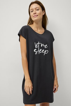 Vrouw draagt een zwarte nachtjapon met korte mouwen met de tekst "laat me slapen" in het wit op de voorkant, staand tegen een effen muur.
