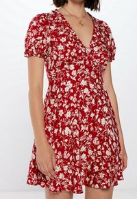 Rotes Blumenkleid mit kurzen, puffartigen Ärmeln und V-Ausschnitt. Verfügt über weiße Blumenmuster, einen ausgestellten Rock und leichten Stoff.