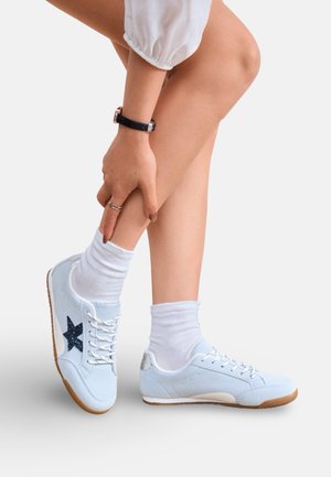Personne portant des baskets bleu clair avec des lacets blancs et des chaussettes, tenant une cheville, montrant les détails latéraux et arrière de la chaussure.