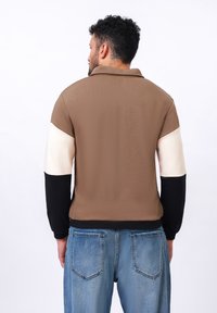 Pull marron avec des manches contrastantes crème et noires, doté d'un col et d'un ourlet côtelé. Texture douce et coupe décontractée.