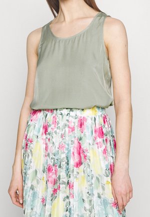Kobieta w zielonym, bezrękawnikowym topie w kolorze szałwii oraz spódnicy z wysokim stanem, plisowanej, z nadrukiem kwiatowym w kolorach różowym, żółtym i zielonym na białym tle.