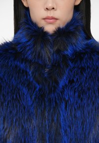 Γυναίκα που φοράει ένα χοντρό, μπλε και μαύρο γούνινο μπουφάν με ψηλό γιακά, απεικονιζόμενη από το κάτω μέρος του προσώπου μέχρι τους ώμους.