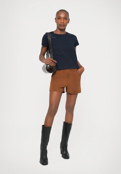 T-shirt à manches courtes bleu marine avec broderie de feuilles, associé à un short en daim marron et des bottes noires montantes, tenant un sac bandoulière rond.