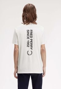 Weißes Baumwoll-T-Shirt mit schwarzem Text auf dem Rücken: "FRED PERRY SEIT 1952" und einem schwarzen Lorbeerkranz-Design. Kurze Ärmel, normale Passform.