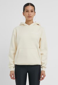 Krämfärgad hoodie med känguruficka, ribbade ärmmuddar och "GOOD KARMA"-logotyp i vitt. Mjuk tyg och avslappnad passform.