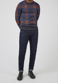 Pull gris avec motif à carreaux bleu et orange, associé à un jean foncé en denim et des baskets noires. Coupe décontractée avec poignets et ourlet côtelés.