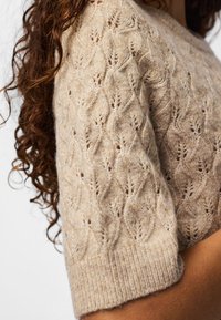 Gros plan sur une manche de pull en maille beige avec un motif de feuille texturé portée par une personne aux longs cheveux bouclés.