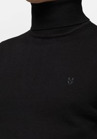 Pull à col roulé noir en tissu côtelé, avec un logo discret sur la poitrine et une coupe ajustée épousant le cou.