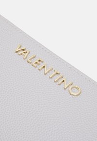 White textured handbag featuring a gold "Valentino" logo. The design includes a smooth, embossed surface and rounded edges.